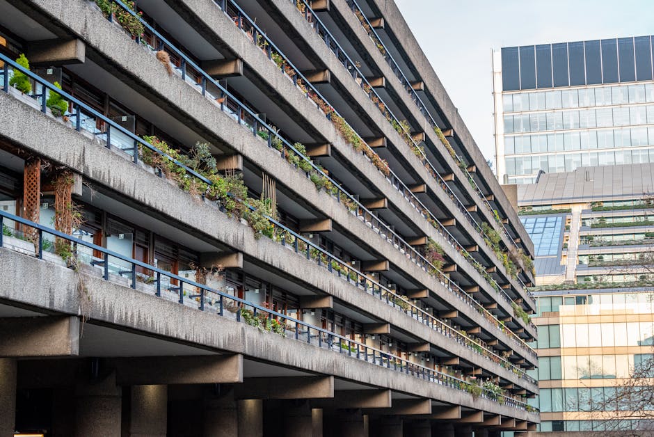 A modern residential building with a Brutalist architectural style, featuring raw concrete surfaces, large rectangular and curved windows, and multiple balconies. In the foreground, there are landscap