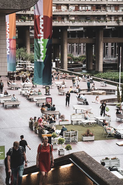 A paved outdoor area adjacent to modern high-rise buildings in an urban setting, featuring a row of wooden benches along a metal railing facing a narrow waterway with reflective surface. The waterway 