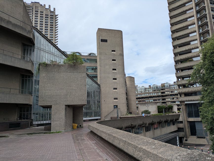 A multi-story urban residential building featuring several balconies with various plants, shrubs, and flower pots. The balconies have metal railings painted in dark blue, and the building's concrete e